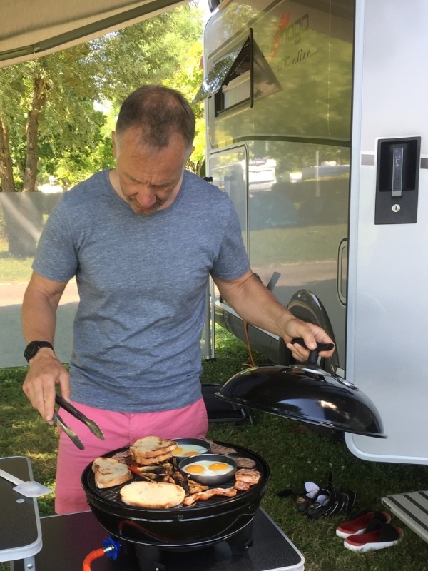 man in grey t-shirt and pink shorts cooking outside a motorhome