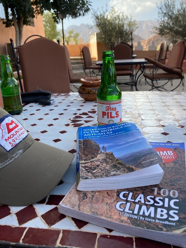 green bottle of Pils and several climbing books on a tiled table with mountains in the background