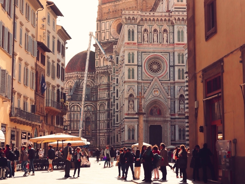 people in a sunny street lined with cafes and a marble Renaissance cathedral in the background