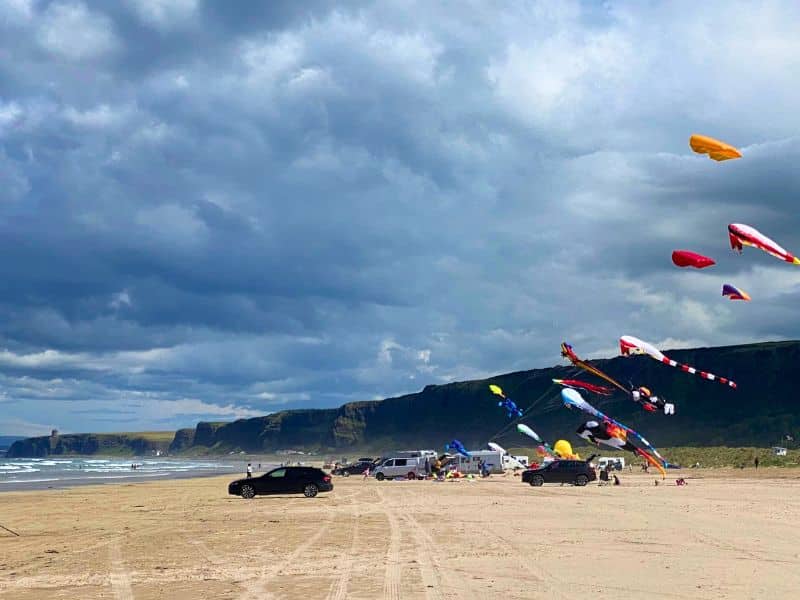 cars and motorhomes parking on a long sandy beach with kites being flown in a stormy sky