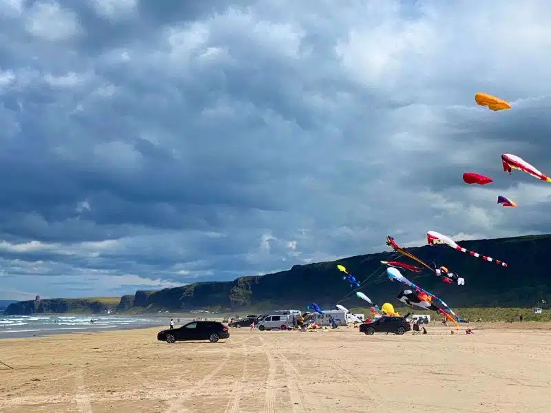 cars and motorhomes parking on a long sandy beach with kites being flown in a stormy sky