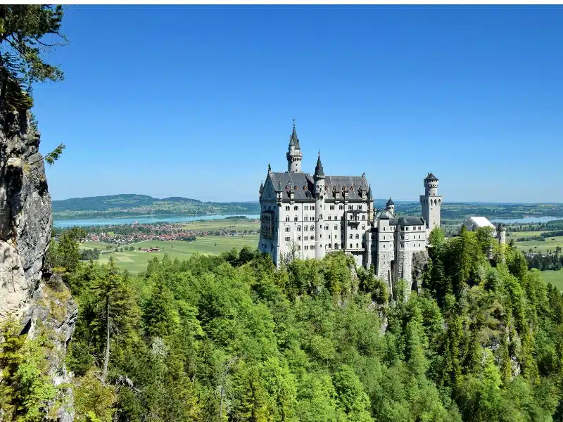 Multi storied white castle in a forest with turrets and far reaching views to a turquoise lake