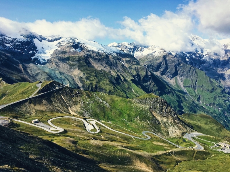 Tarmac road winding through snow capped mountains and alpine meadows