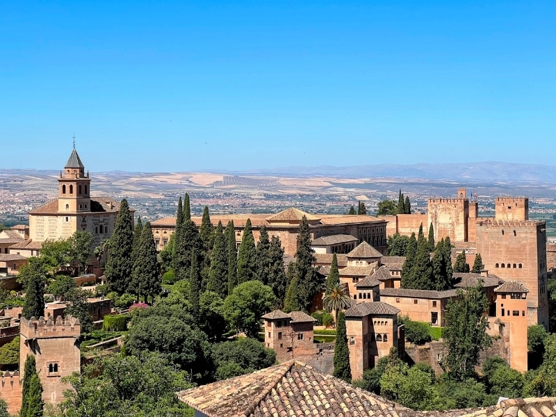 A scenic, slightly elevated view of the Alhambra palace and its surrounding gardens in Granada, Spain, showcasing the historic architecture and lush greenery under a clear blue sky.