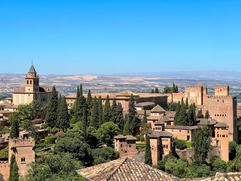 A scenic, slightly elevated view of the Alhambra palace and its surrounding gardens in Granada, Spain, showcasing the historic architecture and lush greenery under a clear blue sky.