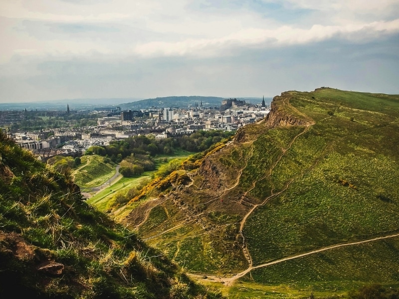 Craggy outcrop with walking trails through grass, overlookig a city with a castle on a hill