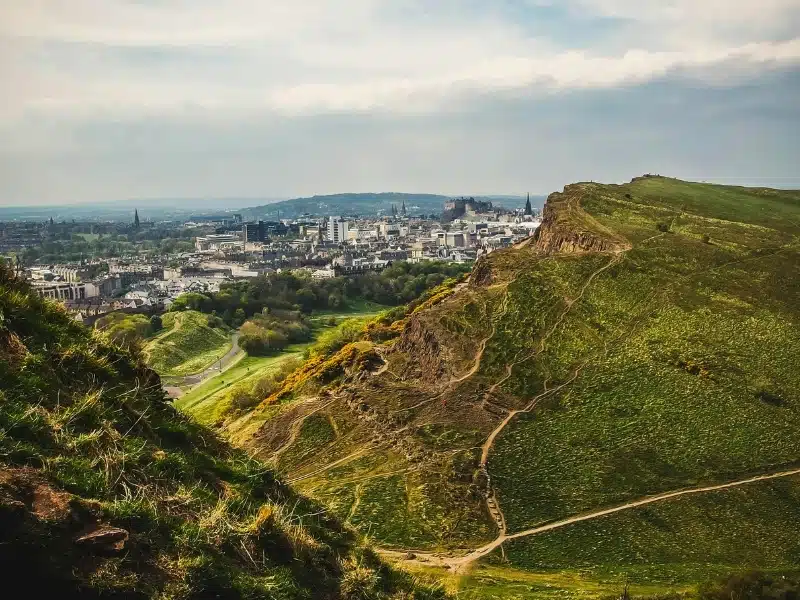 Craggy outcrop with walking trails through grass, overlookig a city with a castle on a hill