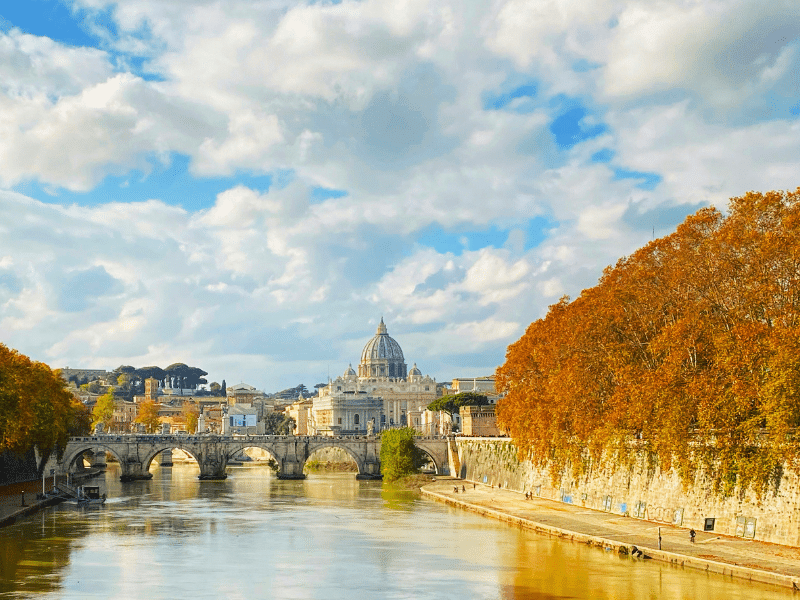 Arched bridge over a still river with vivid orange foliage and a domes church in the background