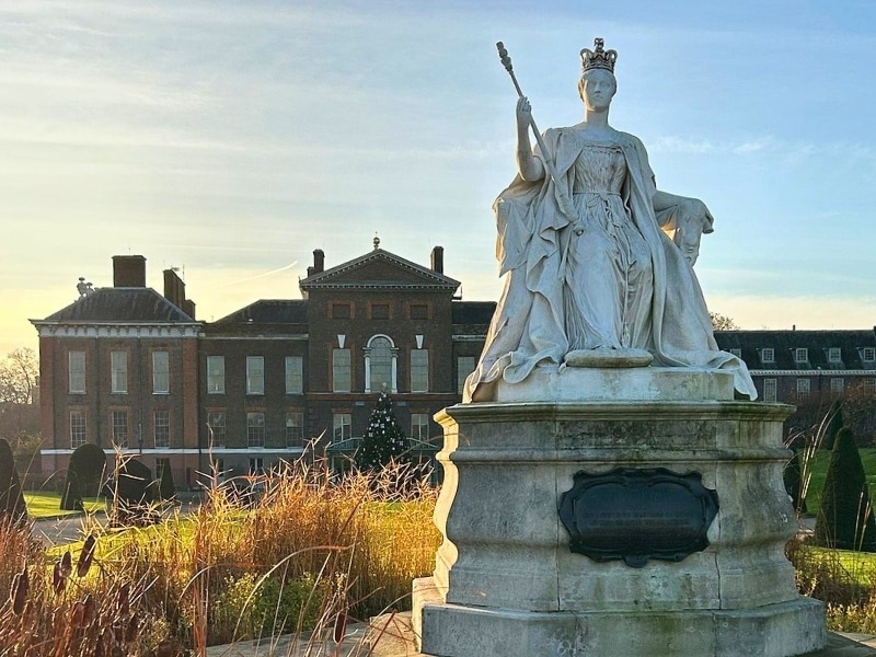 Large white marble statue surrounded by grasses in front of a palace