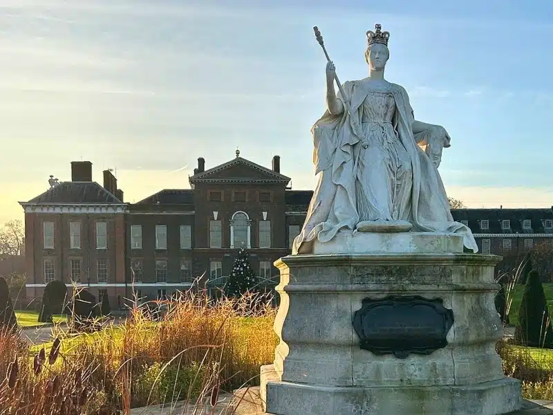 Large white marble statue surrounded by grasses in front of a palace