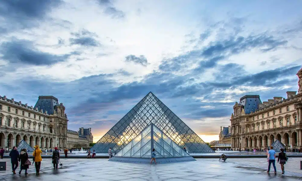 Elegant buildings line a paved square with a double glass pyramid rising from the paving as people walk around it