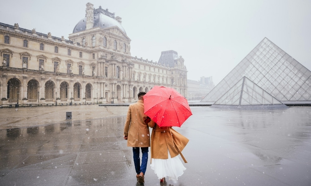 Couple by a large glass pyramid wearing warm coast and carrying a red umberalla