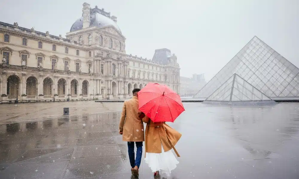 Couple with a red umberalla walking across a large paved area in the rain with a modern glass pyramid and historc buildings surrounding them