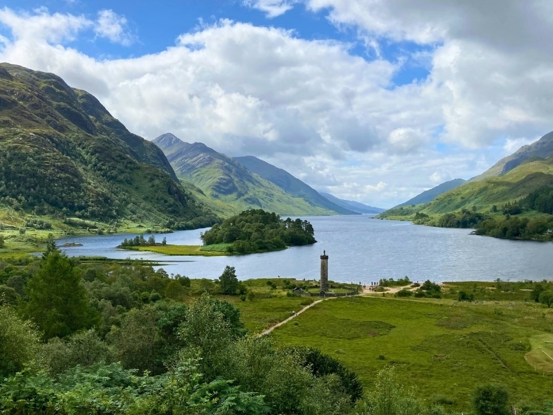 A loch surrounded by mouantsins wiht a tall column memorial by the water