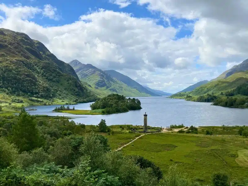 A loch surrounded by mouantsins wiht a tall column memorial by the water