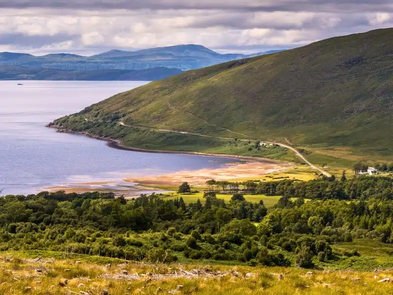 View over a small sandy bay backed by mountains, trees and a few houses