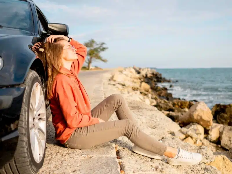 Young woman in an orange hoodie sitting against a car with a sea view