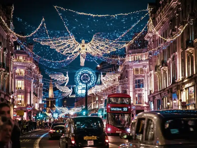 A busy London street with a classis red couble decker bus and black cabs lit for Christmas with lights strung from buildings on either side of the street