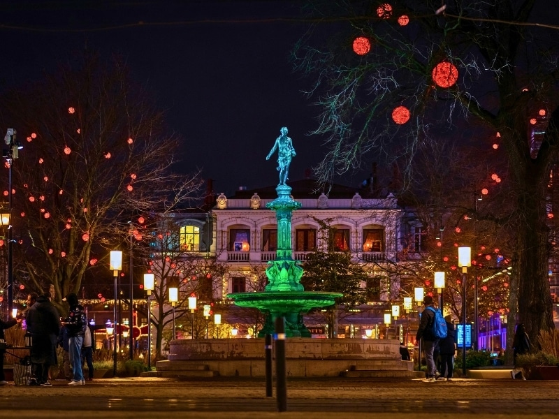 City square at night alight with lanterns and tree decorations with a centrally lit waterfall statue