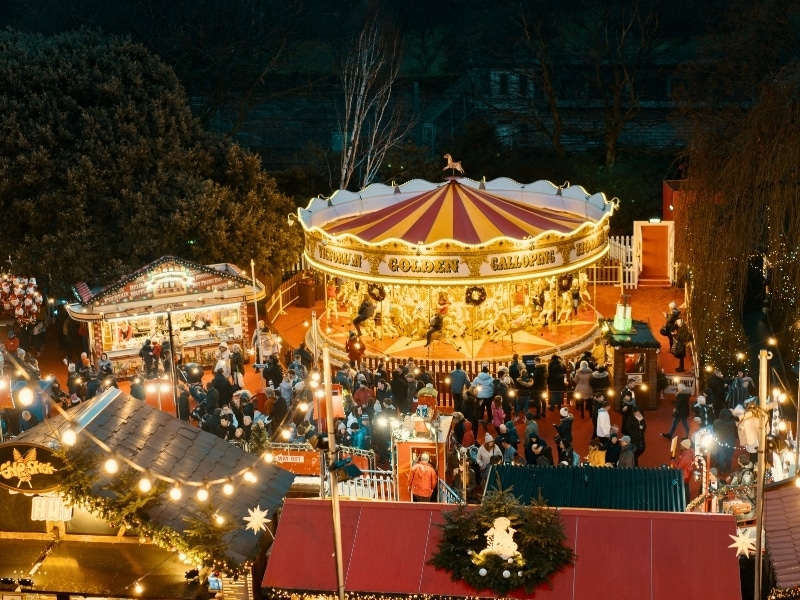 A traditional galloping merry-go-round fairground ride surrouned by people and Christmas Market stalls