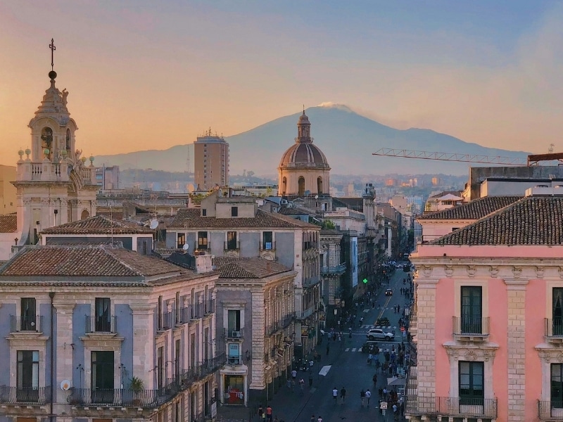 Historic buildings and a domed church line a busy street in the evening with a view of a smoking volcano in the distance