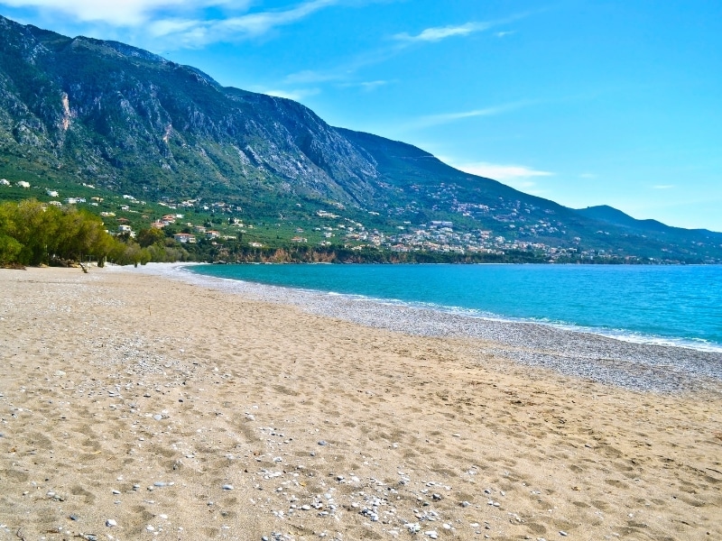 Empty sand and shingle beach lapped by a blue sea wirth houses on the hills in the bakcground