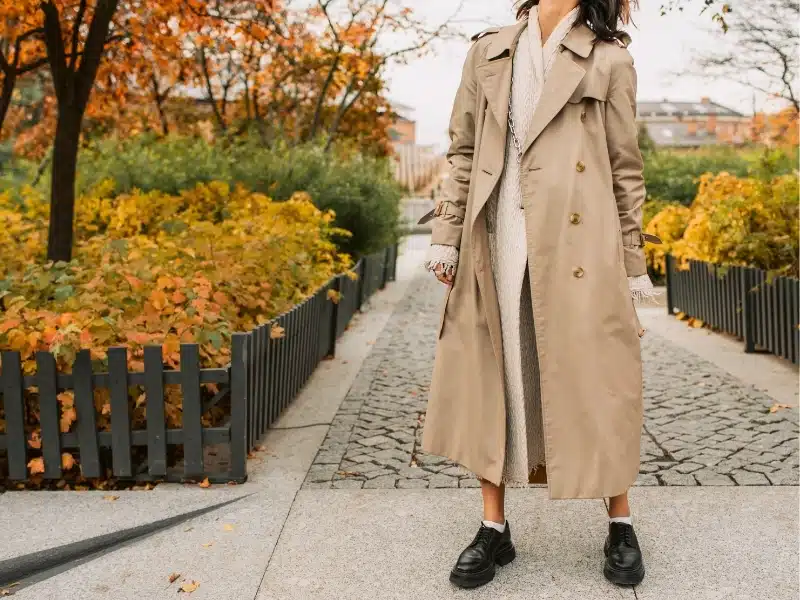 woman wearing a trenchcoat and heavy loafers surrounded by autumn trees in London England