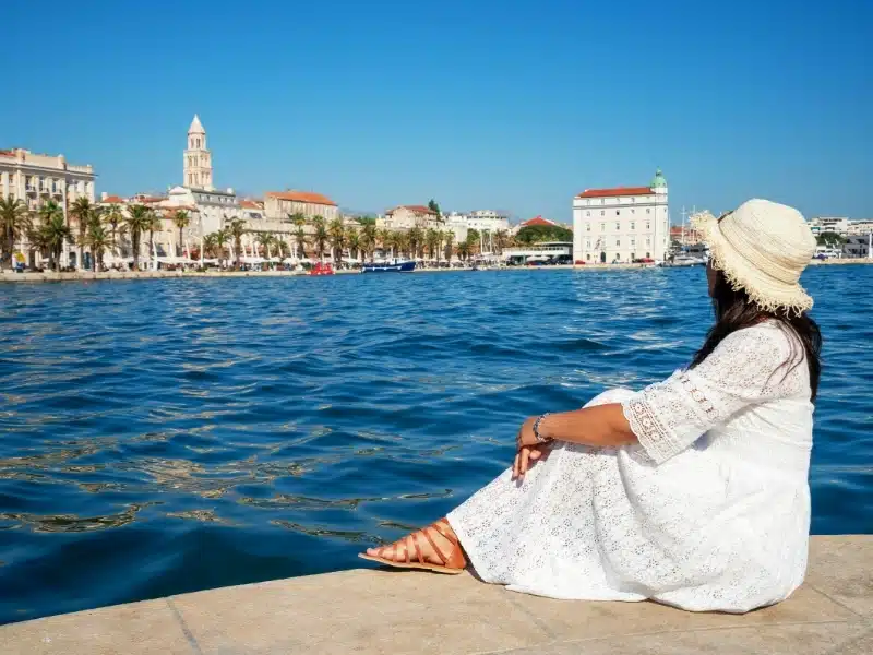 Woman sites by the water in Split Croatia wearing. awhite dress and sandals