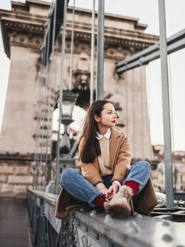Woman sitting on a bridge in Budapest Hungary wearing a camel coat, jeans and boots