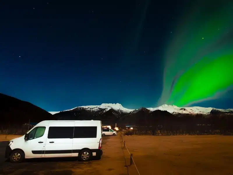 Campervan stand on parking lot in Skaftafell campground with Aurora northern lights at night over snowy mountain peak in Skaftafell national park, Icelandic road trip and transportation. 
