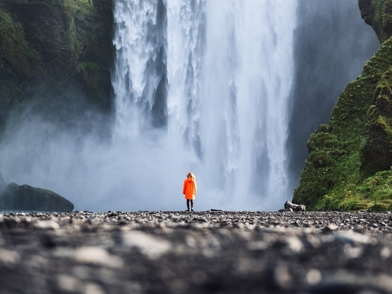 Skogafoss waterfall with a tourist in a waterproof jacker standing in front, over rocky ground