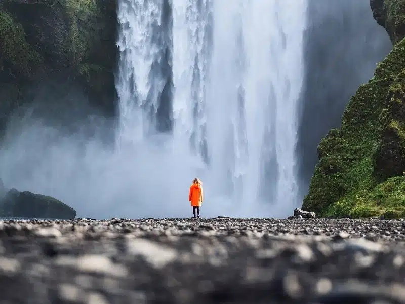 Skogafoss waterfall with a tourist in a waterproof jacker standing in front, over rocky ground