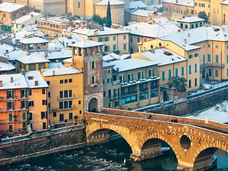 The old town of Verona blanketed by the snow, with the five-arched bridge Ponte Pietra that crosses the river Adige on foreground.