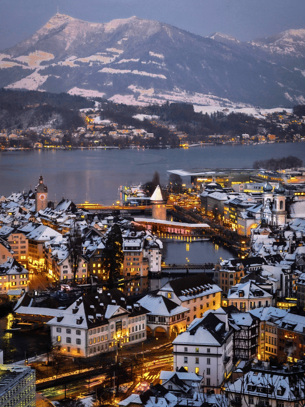 A vertical aerial view of illuminated buildings near the water and a majestic mountain range in Lucerne, Switzerland