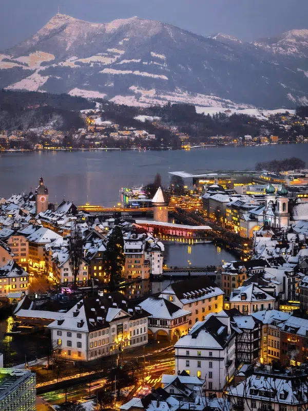 A vertical aerial view of illuminated buildings near the water and a majestic mountain range in Lucerne, Switzerland