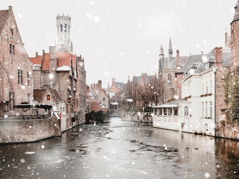 Bruges, Belgium canals view in winter snowfall. Stunning fairy landscape with historical buildings and calm icy water.