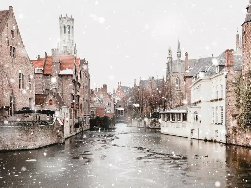 Bruges, Belgium canals view in winter snowfall. Stunning fairy landscape with historical buildings and calm icy water. 