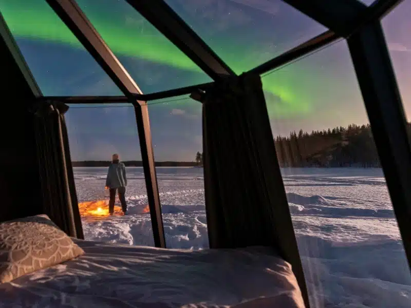 Person with an orange flashlight admiring the norther lights outside a glass igloo in a snow field