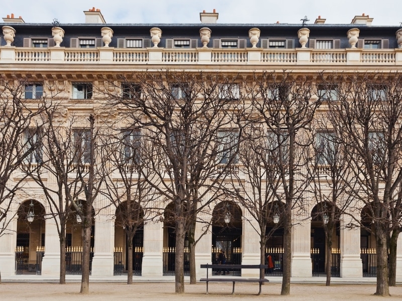 Bare trees line an avenue with an arched portico behind them