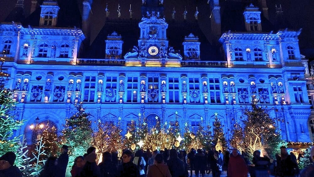 French Renaissance town hall bathed in blue light with many Christmas trees and people at night