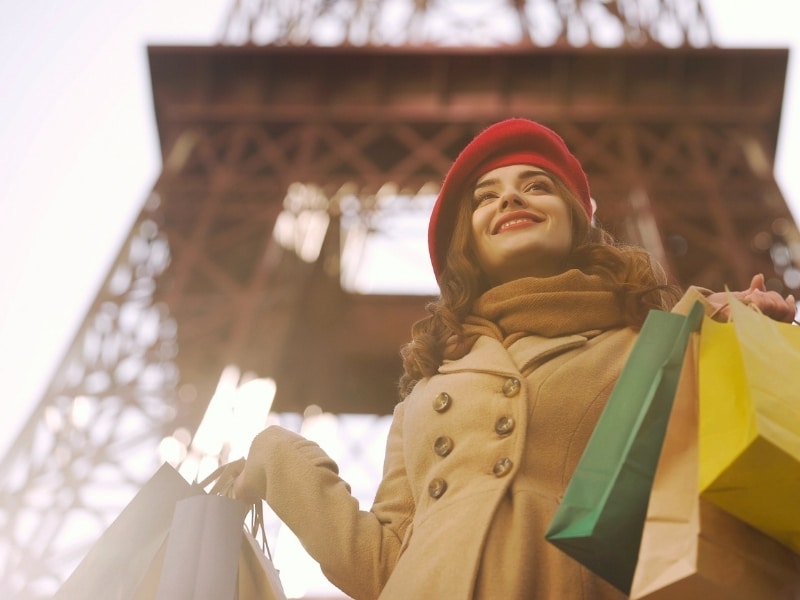Woman in a camel coat and red beret carrying shopping bags in front of the Eiffel Tower in Paris