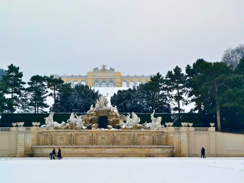 Large ornate and gildedn fountain without water in a snowy setting with a grand building on a hill behind