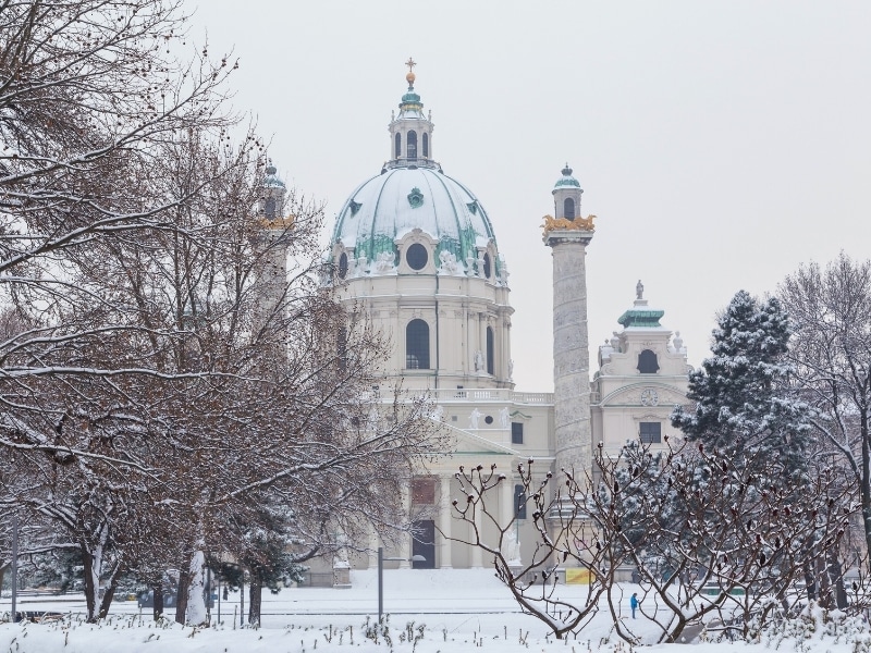 The front of Karlskirche in central Vienna during the winter. Lots of snow and trees can be seen.