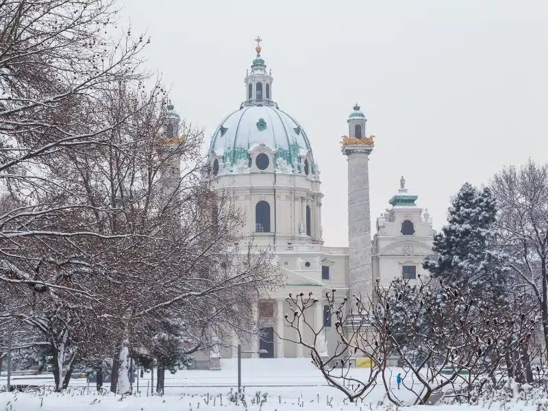 The front of Karlskirche in central Vienna during the winter. Lots of snow and trees can be seen.