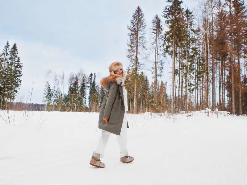 Girl walking in winter snow meadow, wearing stylish fashion outfit khaki parka coat jacket, fur hat and moon boots, full length, looking back. Happy smiling woman hiking in nature
