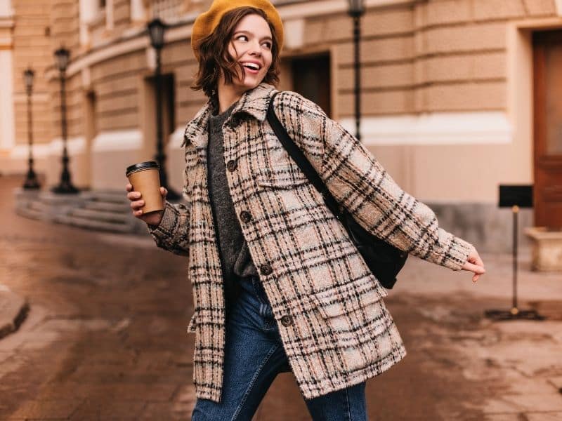 Attractive woman with short hair looks back with smile. Girl in beige coat walks through city with glass of coffee