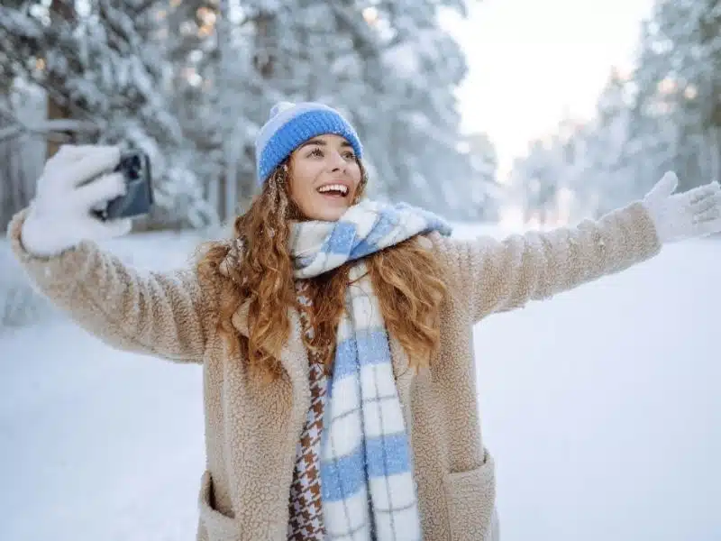 Happy woman in a bright hat and scarf with a phone in her hands takes a selfie in a sunny winter snowy forest. 