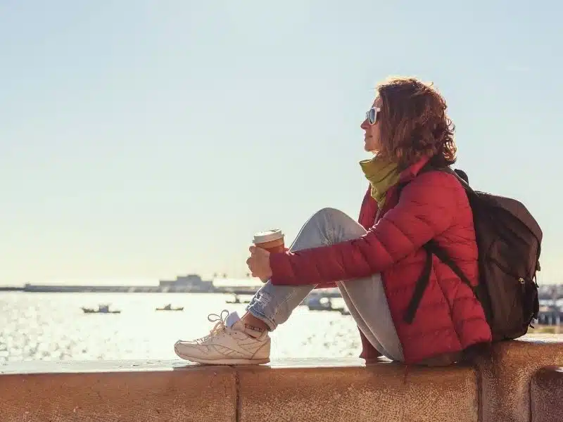 Beautiful young woman in jacket and scarf sits with takeaway coffee overlooking the sea in windy weather