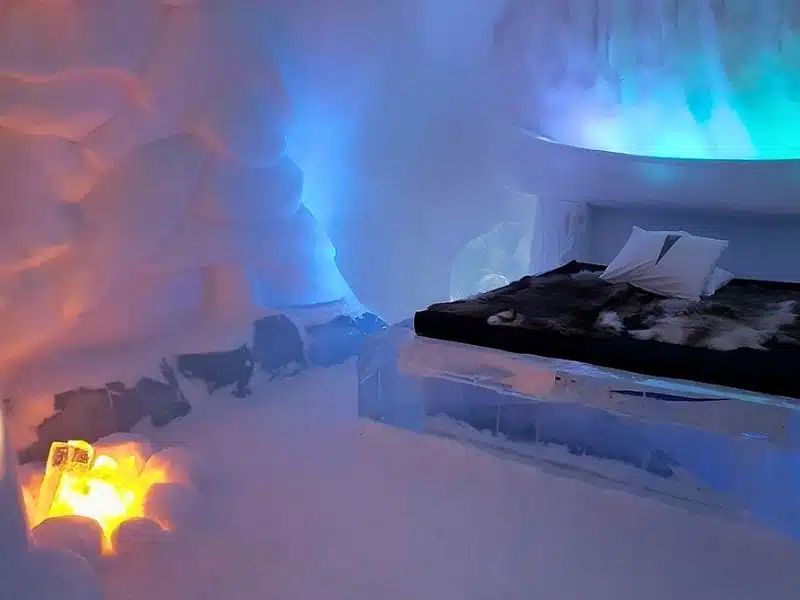 bed made on ice shelf in an igloo stlye room, covered with furs abd a fire in the corner