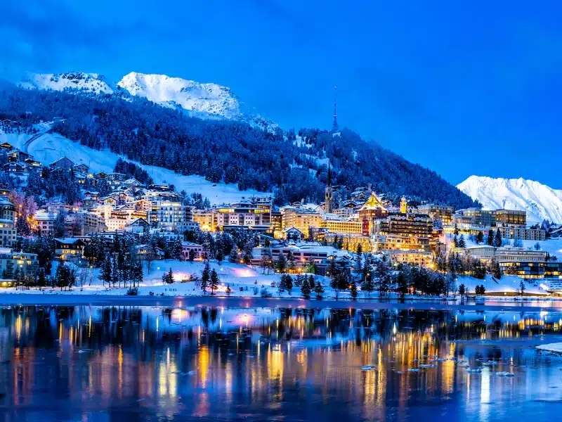 View of beautiful night lights of St. Moritz town in Switzerland at night in winter, with reflection from the lake and snow mountains in backgrouind
